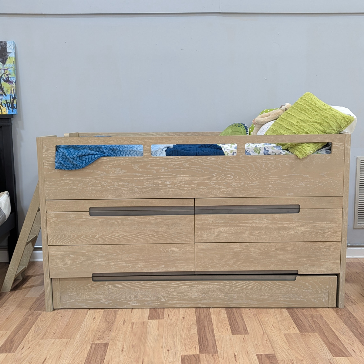 A light oak wood bed frame with a storage drawer unit underneath, styled with bedding and pillows, set against a plain gray wall and wood floor.