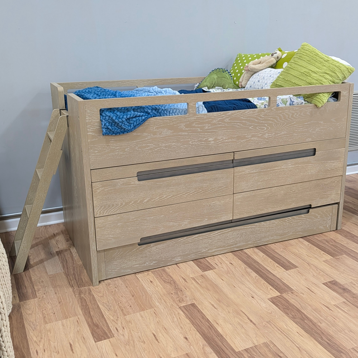 A light oak wood twin loft bed with built-in drawers and a short ladder, styled with blue and green bedding and pillows, set against a gray wall and wood-look floor.
